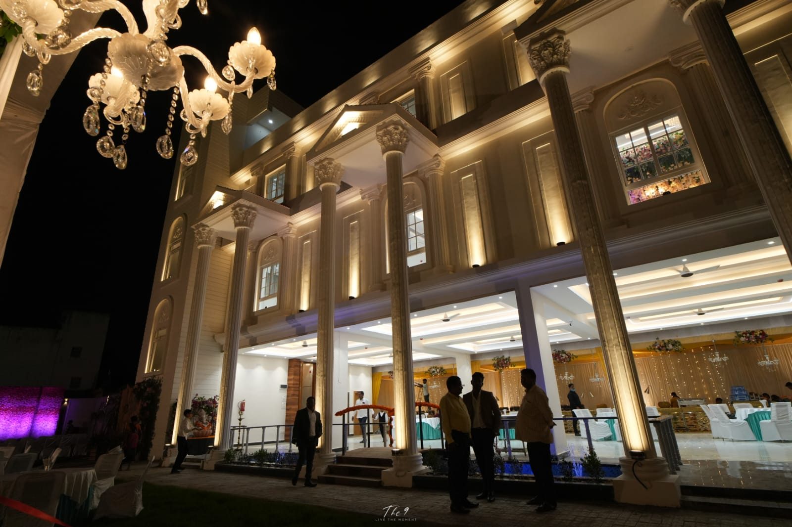 Aarambh Banquet façade lit with warm uplights at night, a crystal chandelier in the foreground