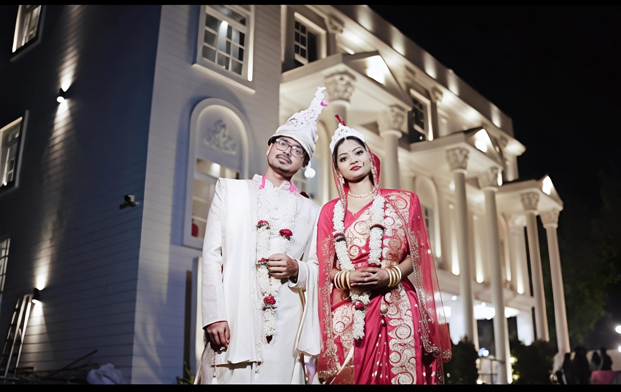A bride and groom in front of the lit Aarambh Banquet façade